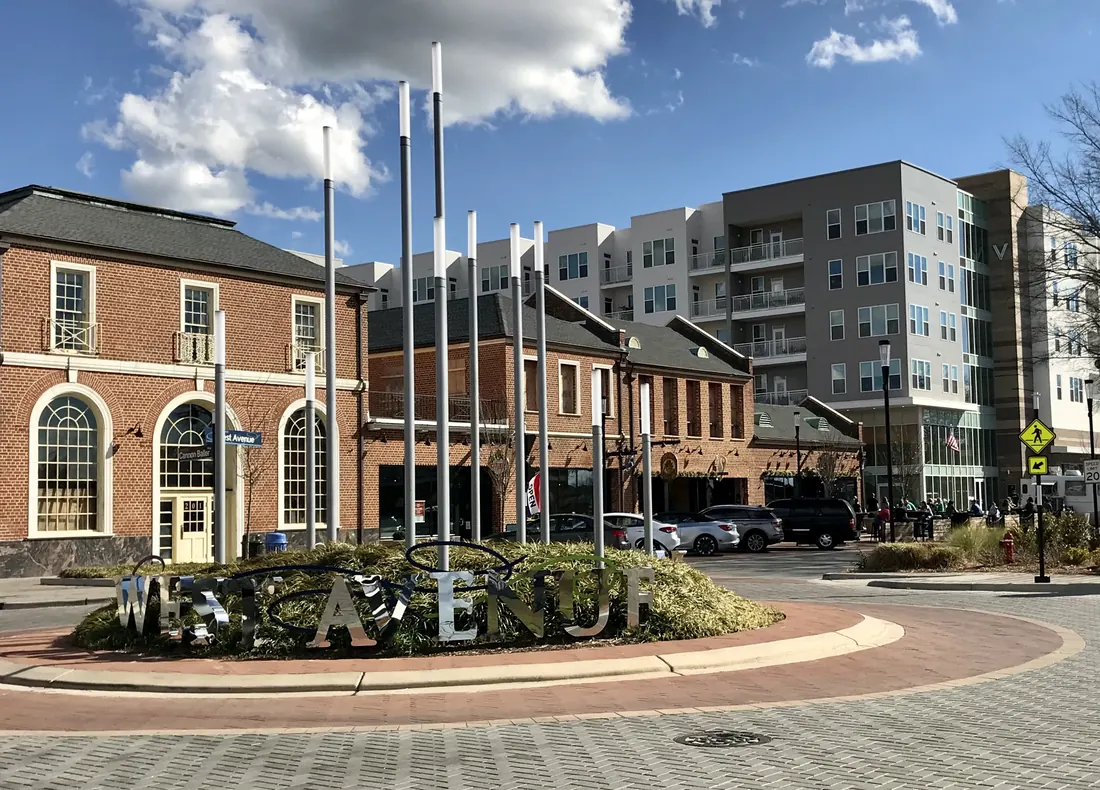 Chrome letters spelling West Avenue in a planted circle in downtown Kannapolis, with red-brick buildings, tall light poles and a new apartment building under a blue sky